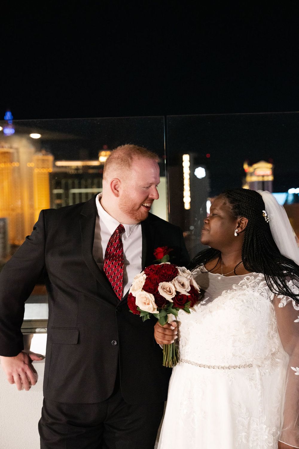 Interracial couple in wedding attire smiling at each other on a rooftop with the Las Vegas Strip at night.