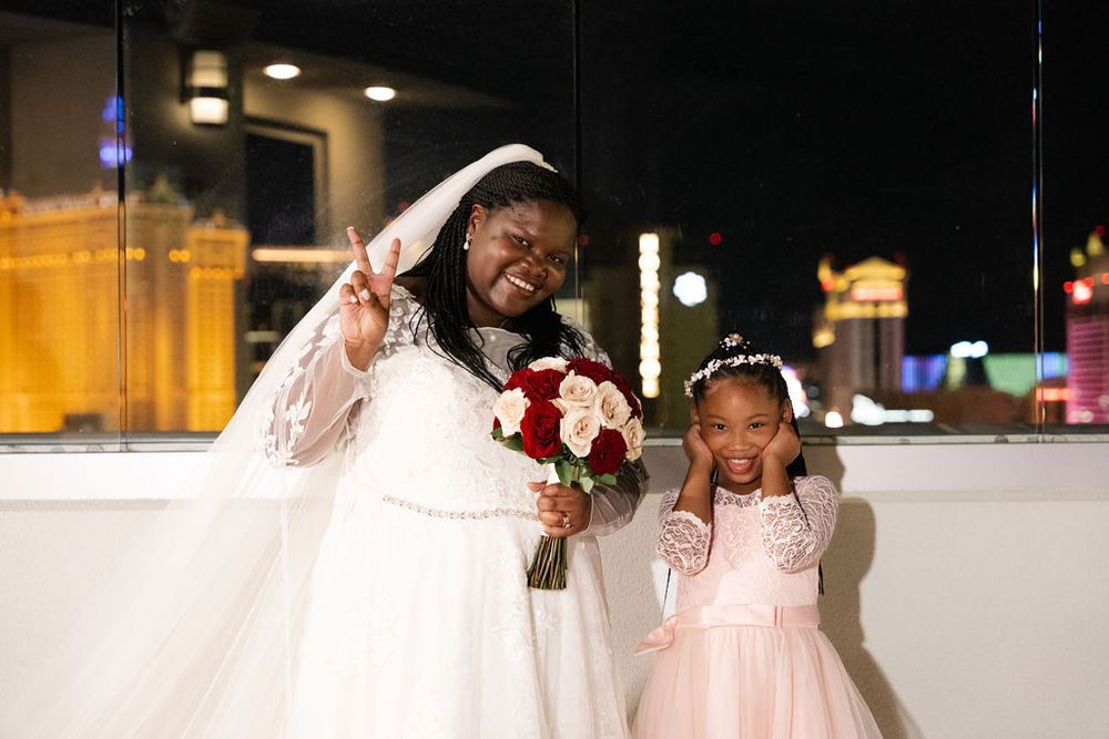 Bride in white gown and veil poses with young flower girl in pink dress, Las Vegas skyline at night.