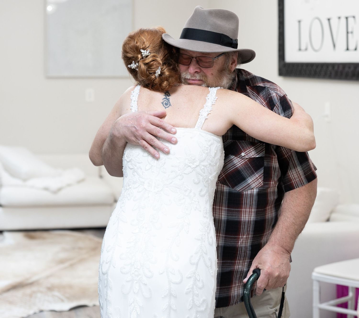 Relative hugging bride in white lace wedding dress, emotional moment, man wearing plaid shirt and brown fedora hat.