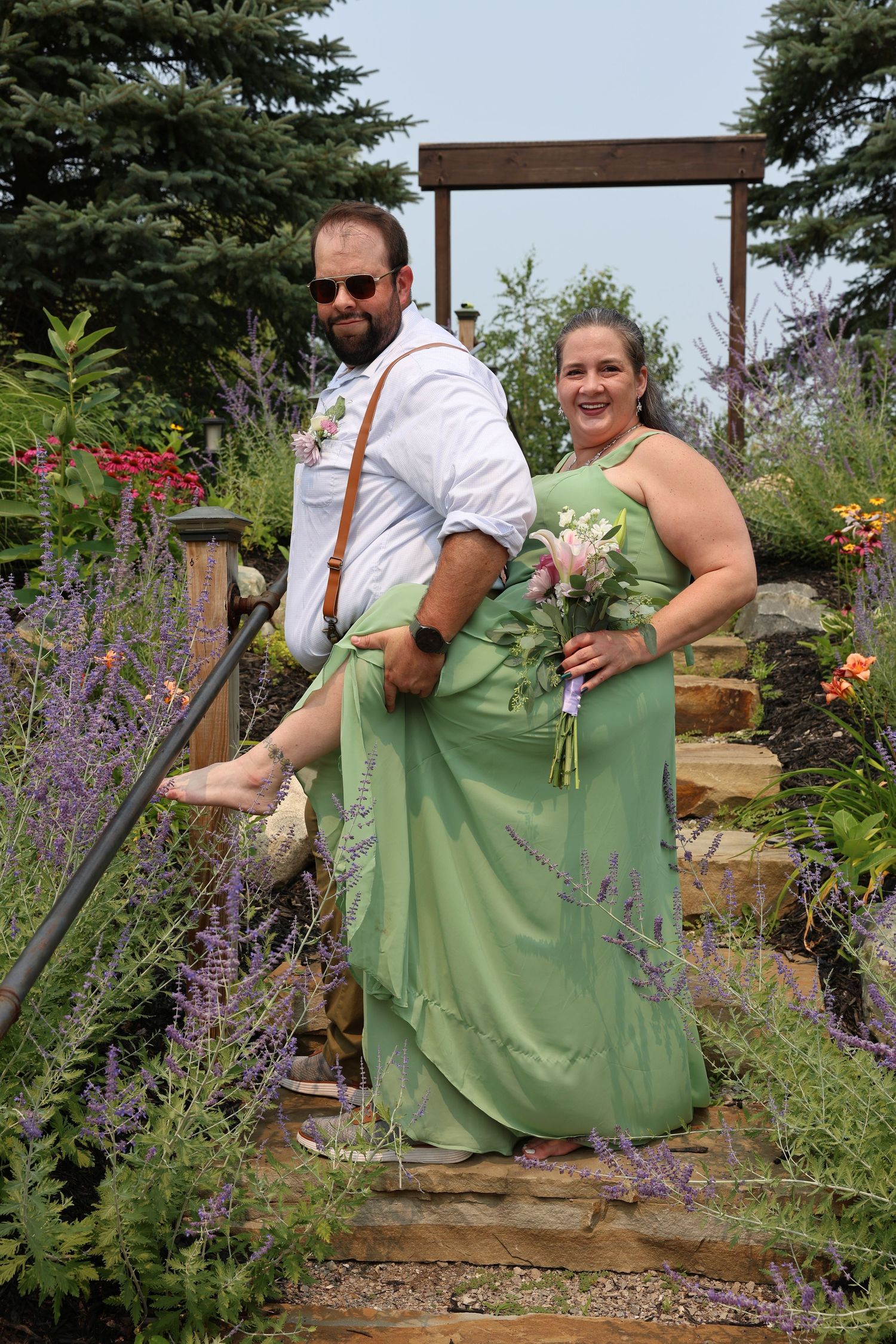 A man and woman in wedding attire pose on garden steps, she holds a bouquet in a green gown, he wears suspenders.