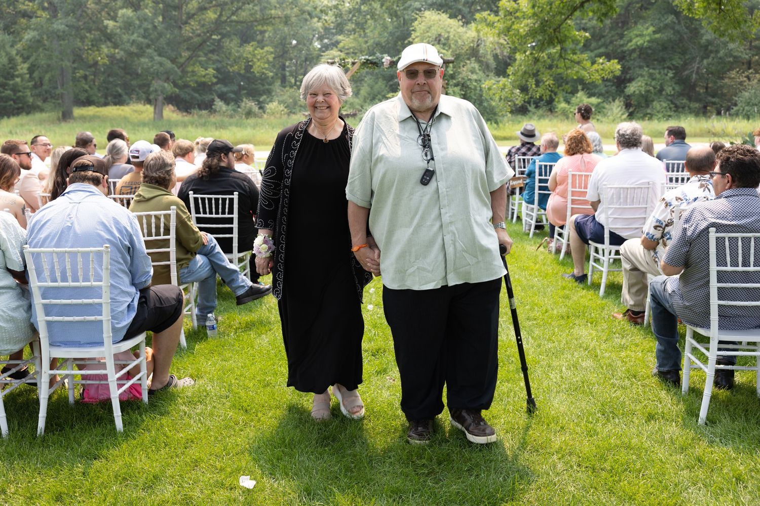 Elderly couple walking down outdoor wedding aisle on green lawn, surrounded by seated guests on white chairs.