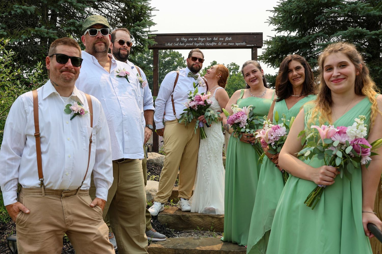 Wedding party in green bridesmaid dresses and tan suits pose outdoors under a wooden arch with floral bouquets.