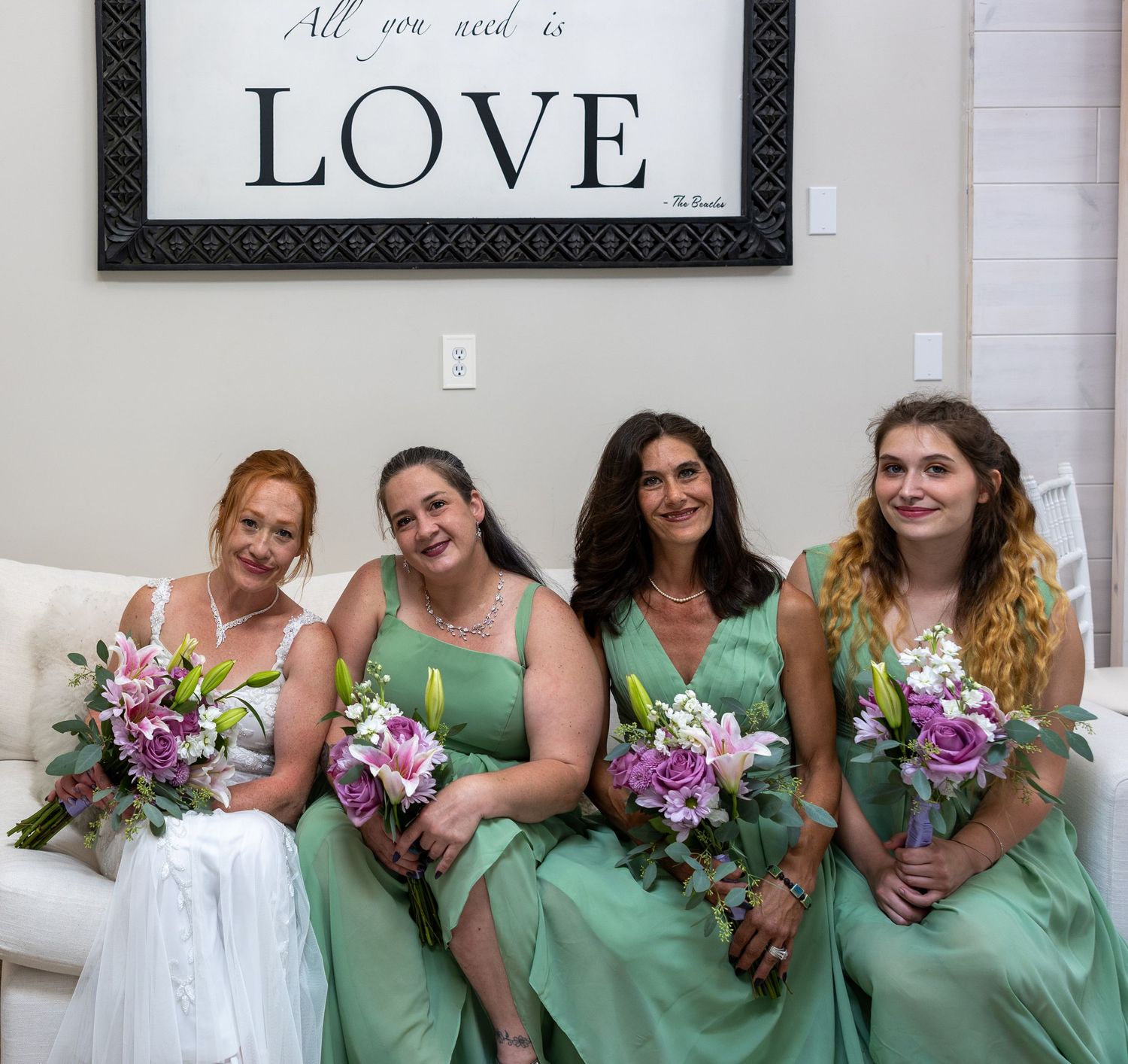 Bride and three bridesmaids in mint green dresses holding purple floral bouquets seated together beneath a Love sign.