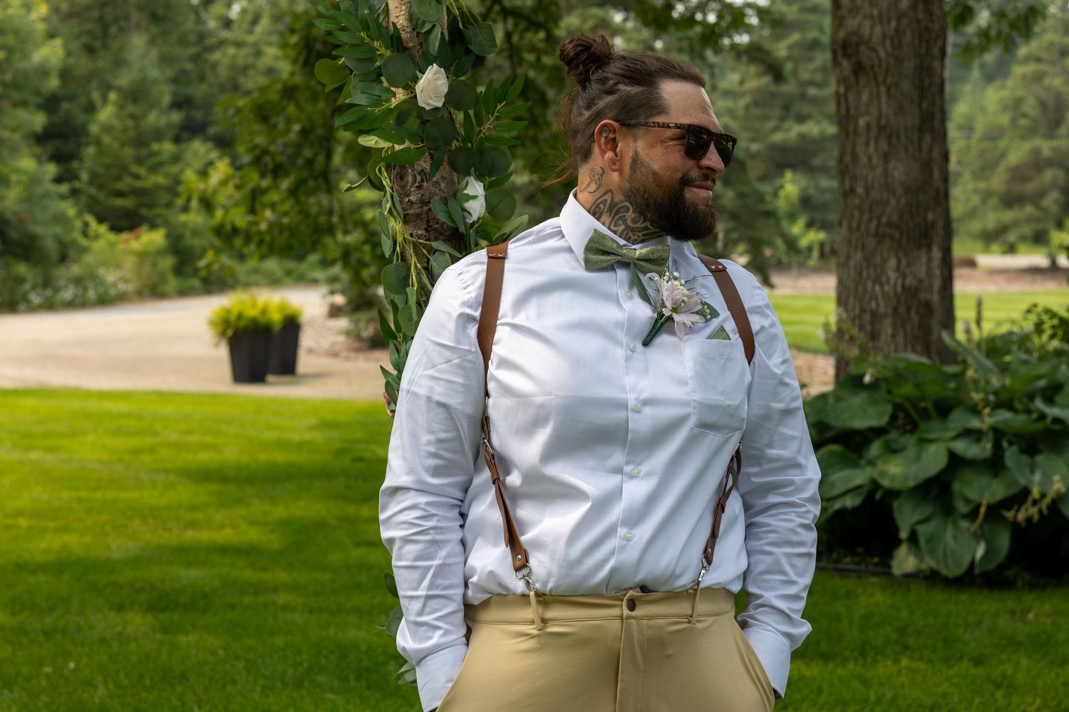 Stylish groom in white shirt, suspenders, and bow tie posing outdoors at a wedding venue.