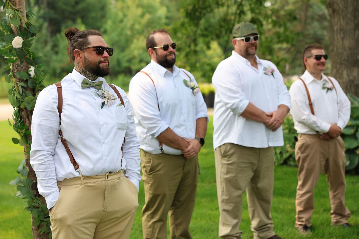 Emotional groom with groomsmen in white shirts, khaki pants, and suspenders stand outdoors at a wedding ceremony on a sunny day.