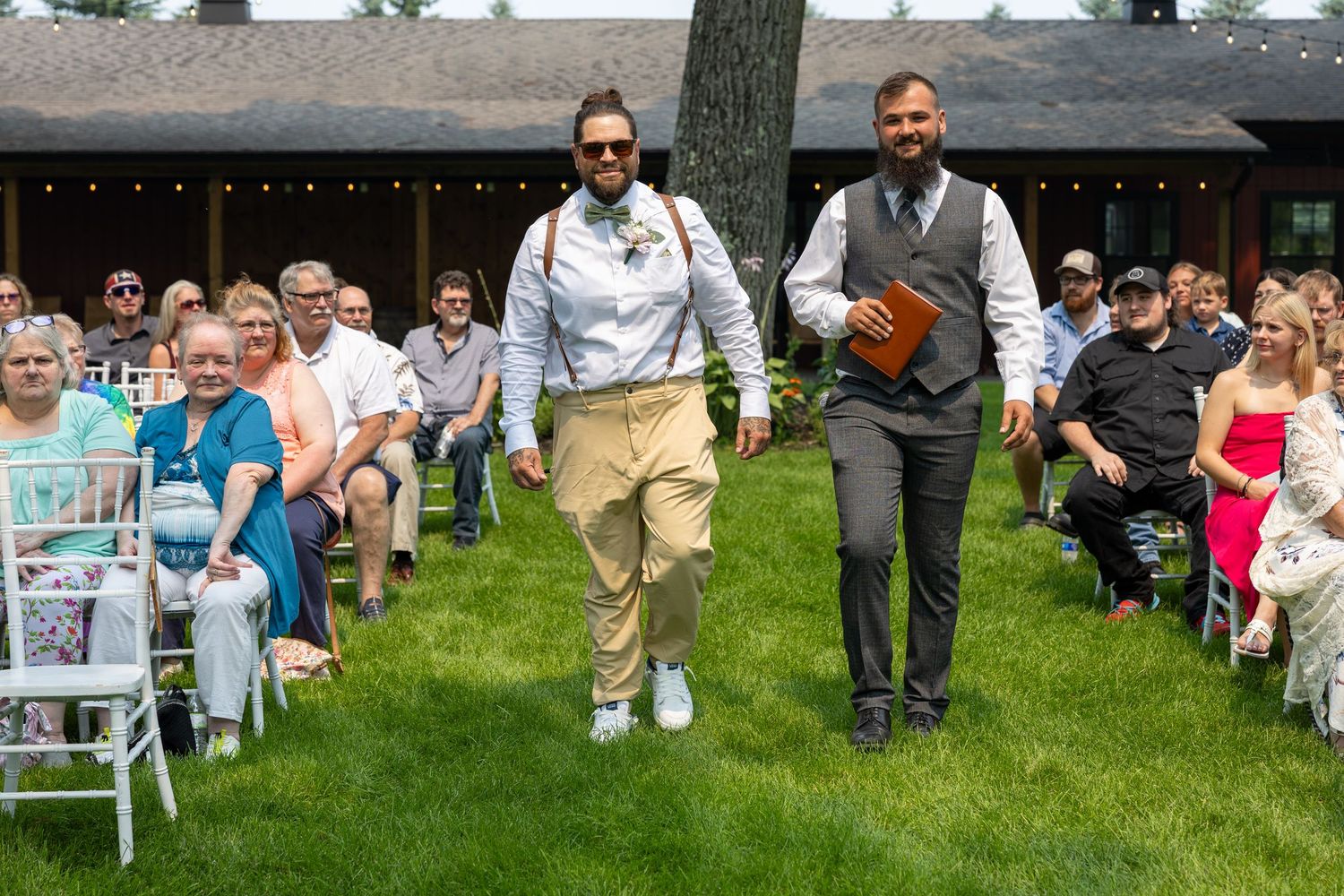 Groom and officiant walking down the aisle at an outdoor wedding ceremony with seated guests on a sunny day.