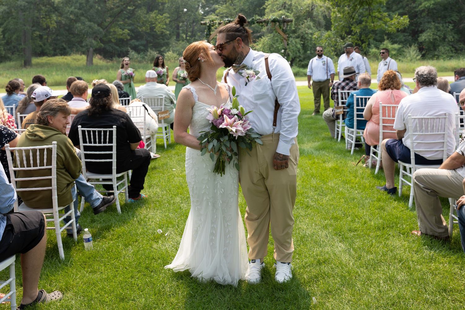 Newlywed couple sharing a kiss at outdoor wedding ceremony on green lawn with guests seated in white chairs.