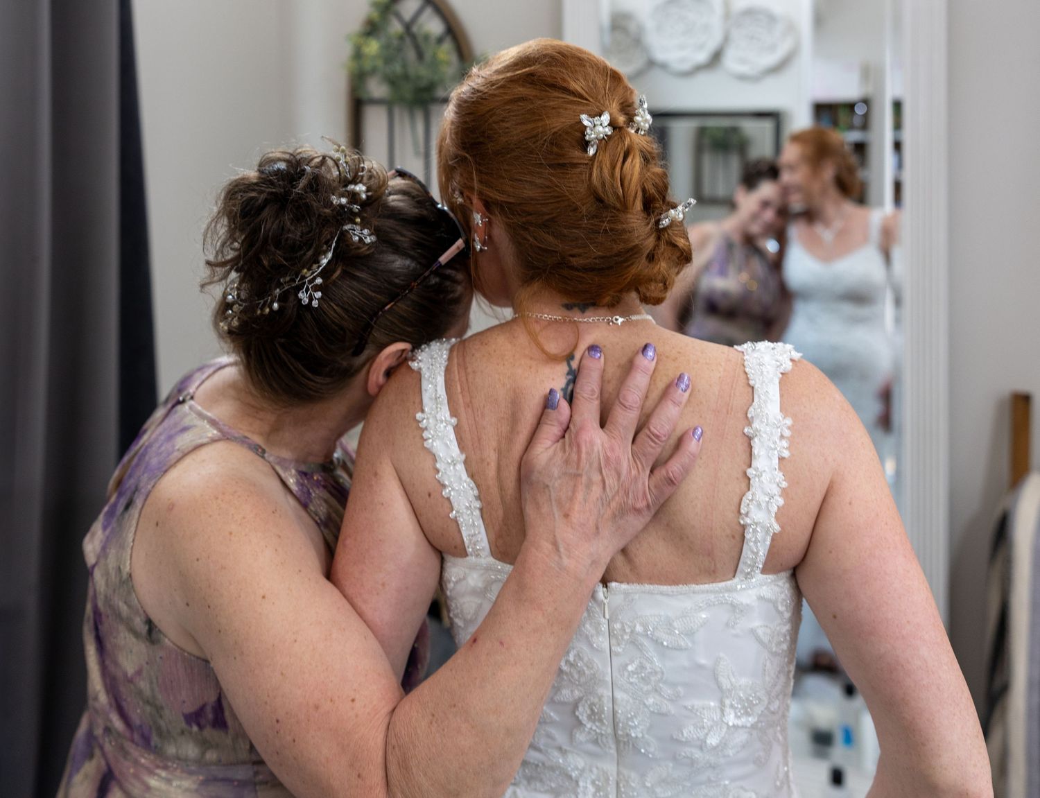 A bride's mom helps a red-haired bride fasten her white wedding dress, with a mirror reflection visible in the background.
