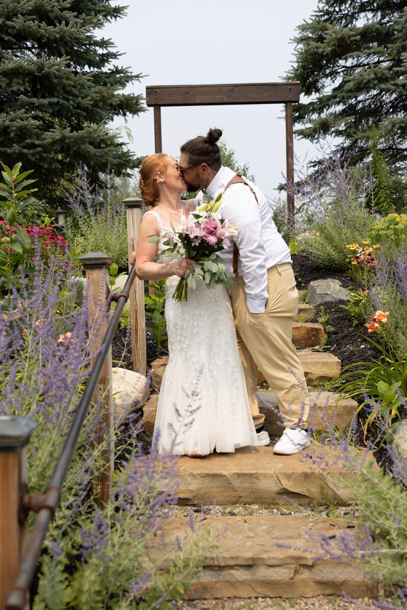 Bride and groom share a kiss on stone steps surrounded by lavender and colorful garden flowers under a wooden arbor.