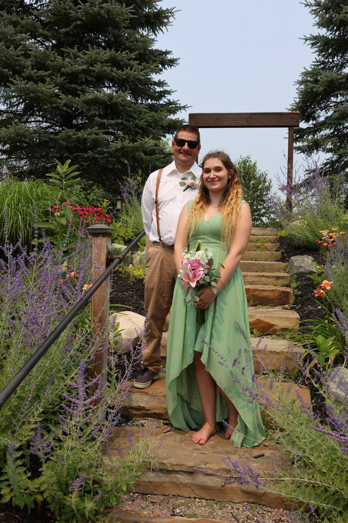 Couple in wedding attire on stone steps surrounded by lavender and flowers, woman in green dress holding pink bouquet.