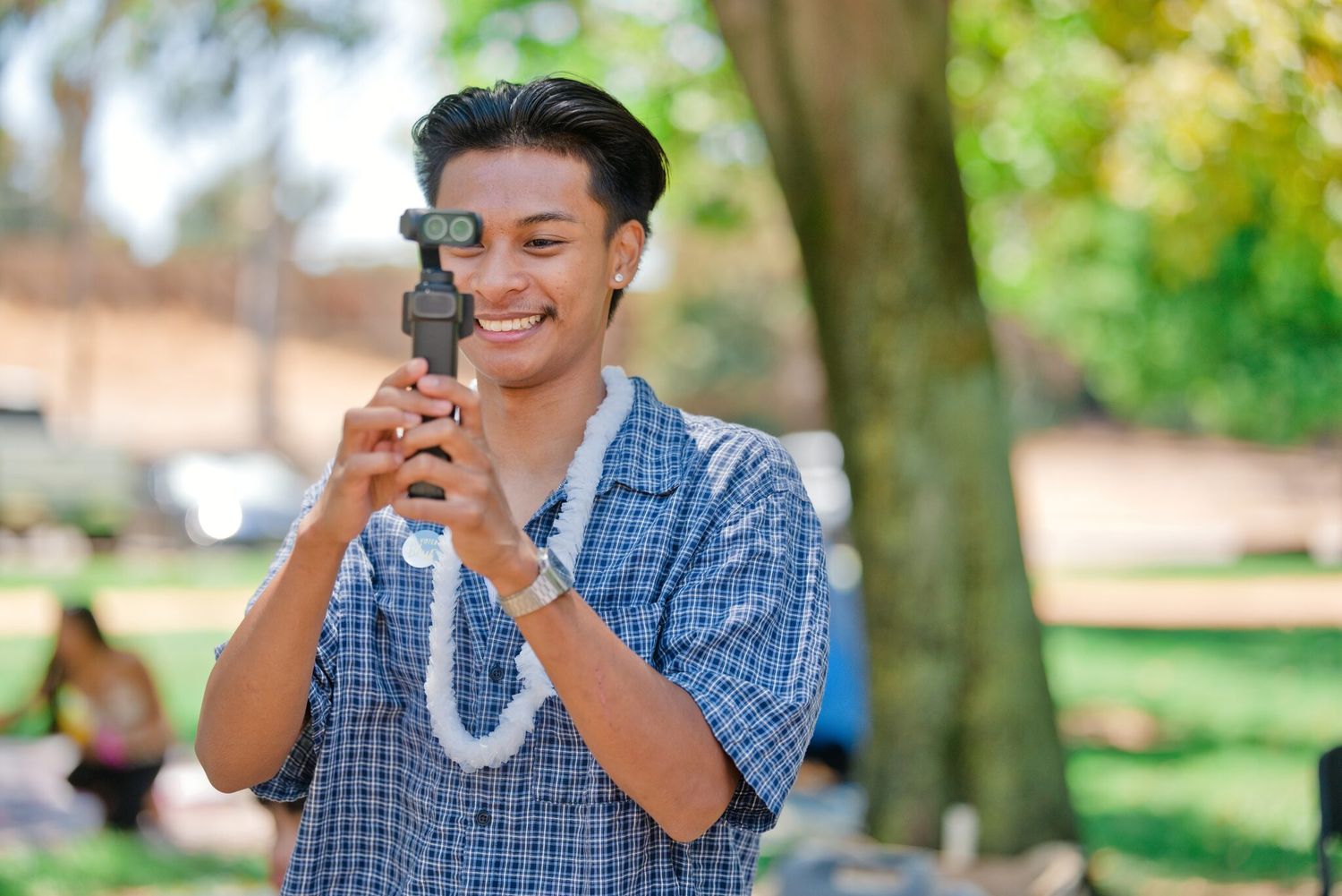 Smiling young man holding a camera gimbal stabilizer outdoors in a sunny park setting.
