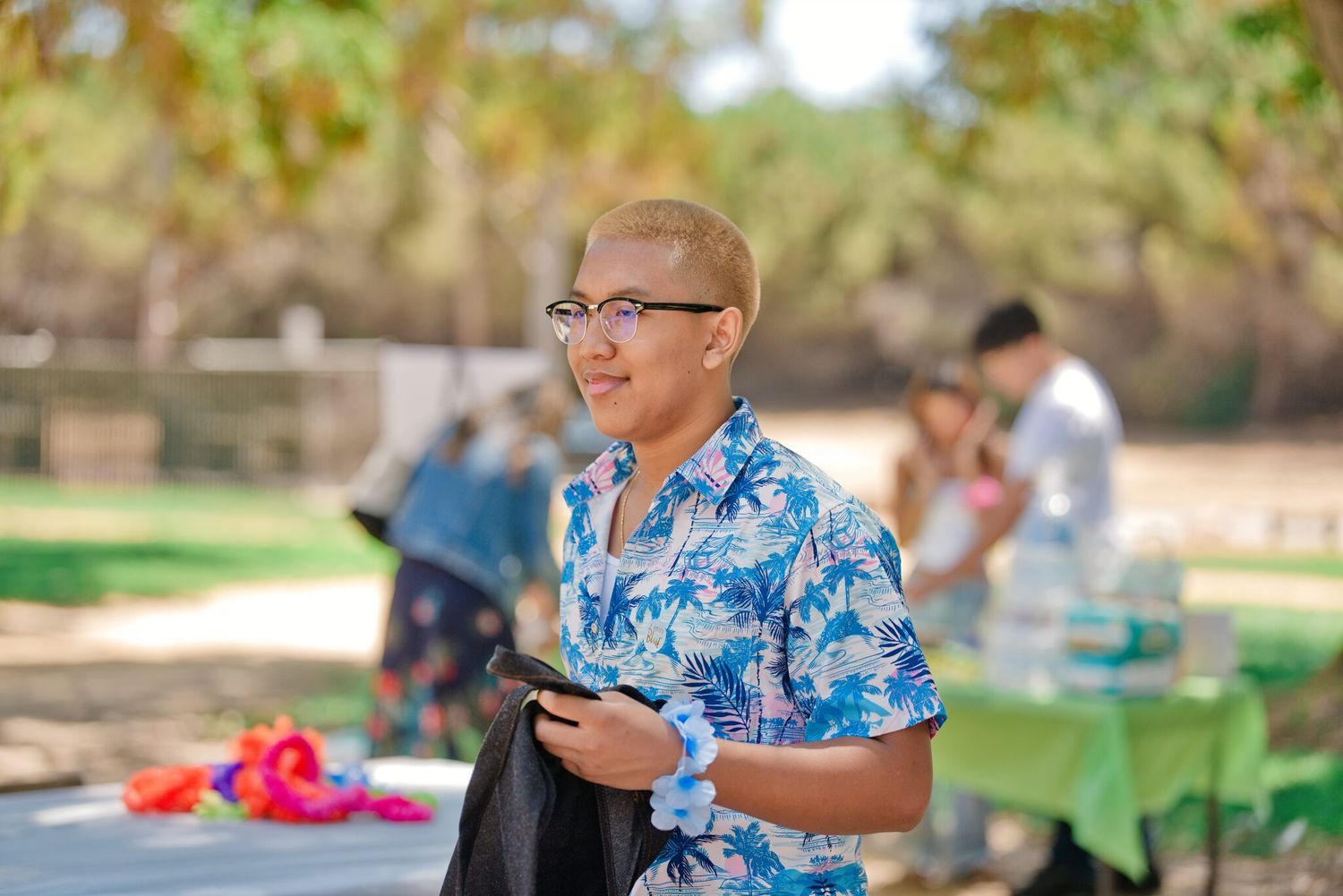 Young person in floral shirt holding phone smiles outdoors at a sunny park gathering.