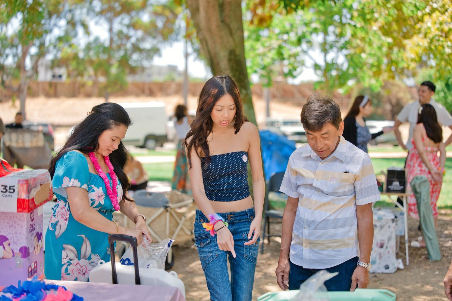 Three people browse colorful items on tables at an outdoor park event on a sunny day.