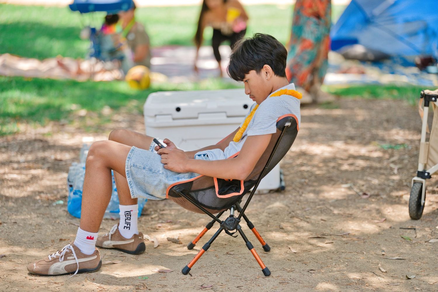 Young boy sitting in a folding camp chair using a smartphone at an outdoor park gathering.