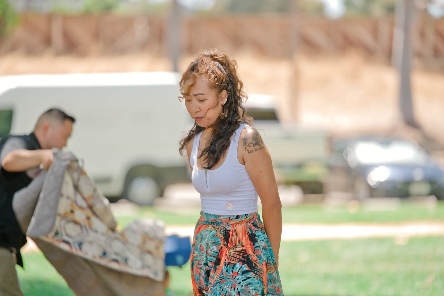 Woman in white tank top and colorful skirt with tattoos standing outdoors in a sunny park setting.