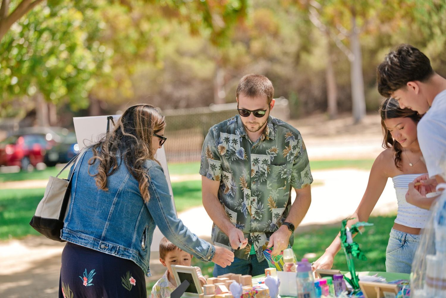 A group of people gather around a table at an outdoor park event on a sunny day.
