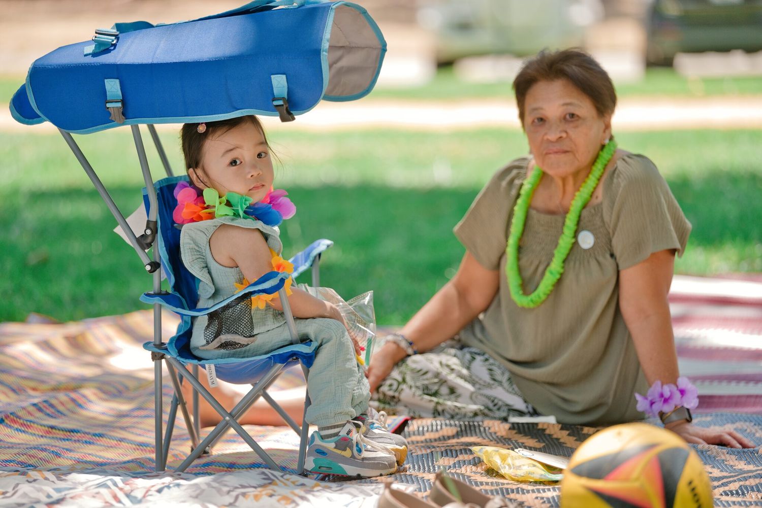 Elderly woman and toddler sitting on blanket in park, child in blue camping chair with canopy, woman wearing green lei.