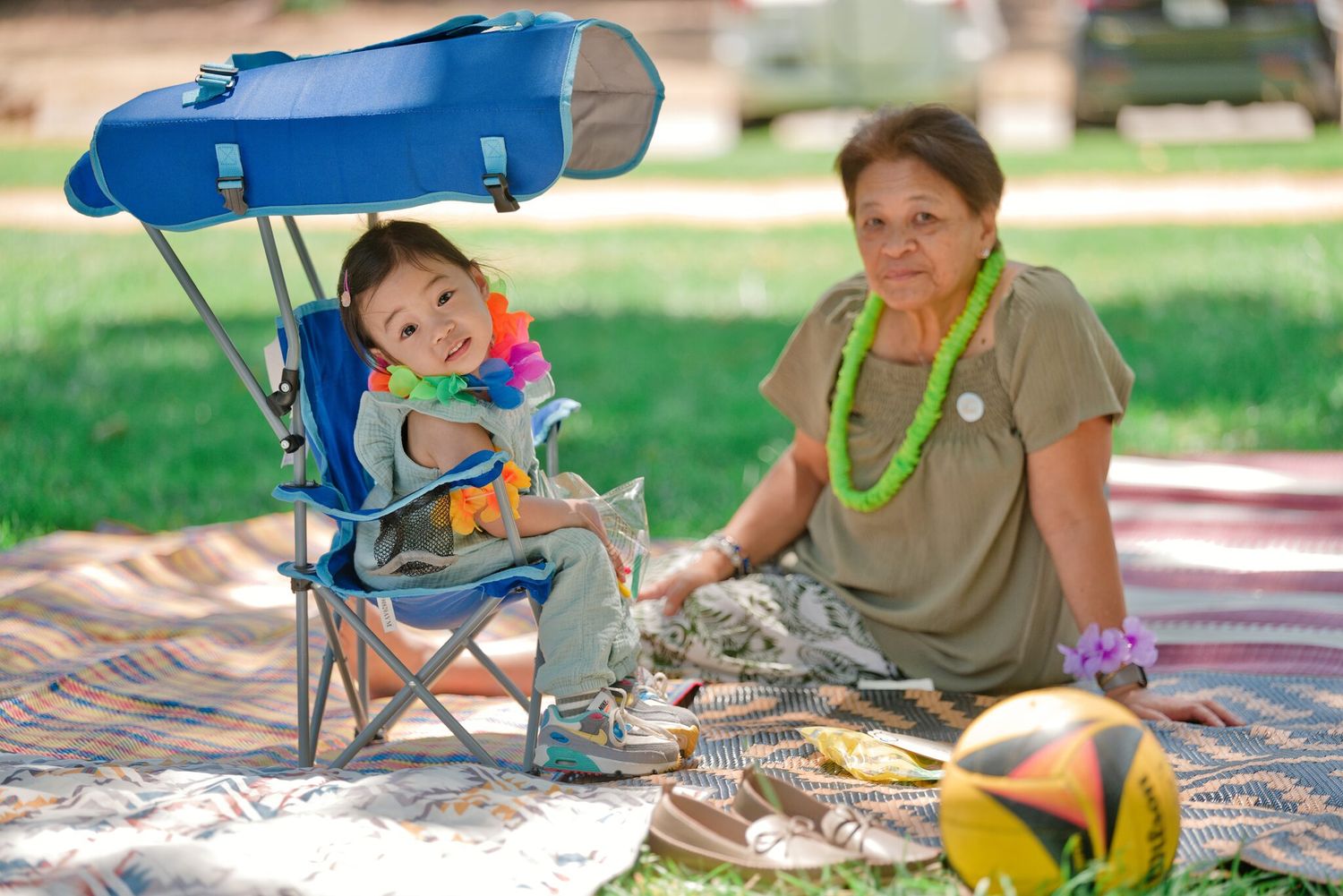 Baby in blue camping chair with canopy beside woman wearing lei on picnic blanket in park.