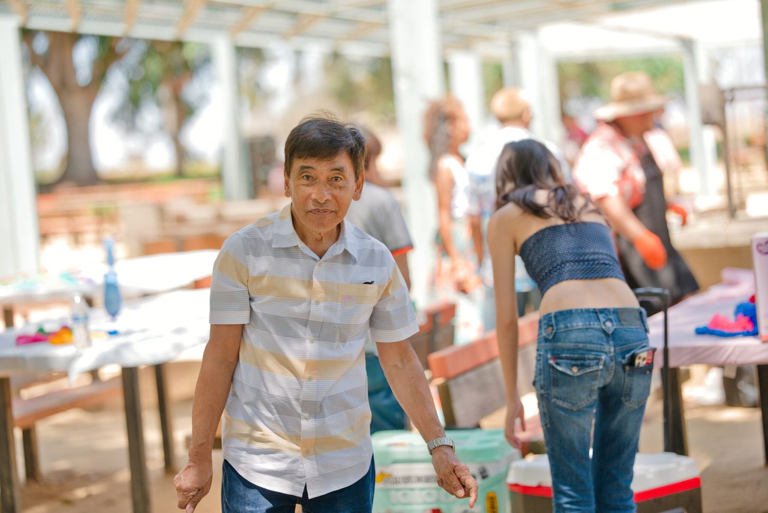 A man in a striped shirt walks through an outdoor gathering area with people and tables in the background.