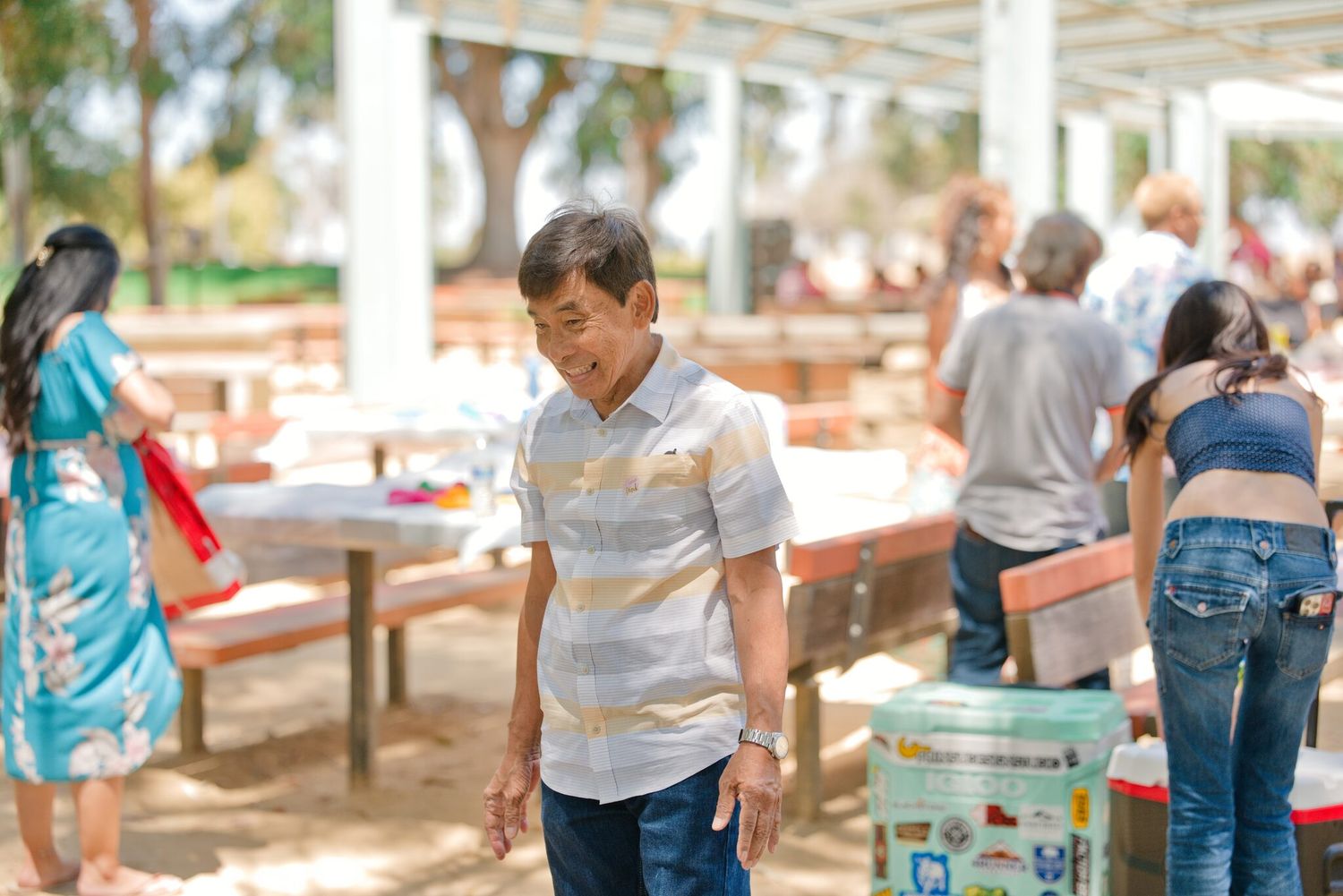 A young boy in a striped shirt stands outdoors at a casual gathering with people and picnic tables in the background.
