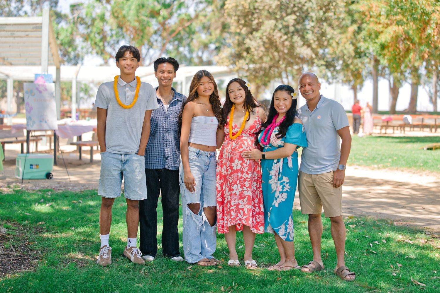 A joyful family of six poses outdoors at a sunny park celebration, some wearing colorful leis.