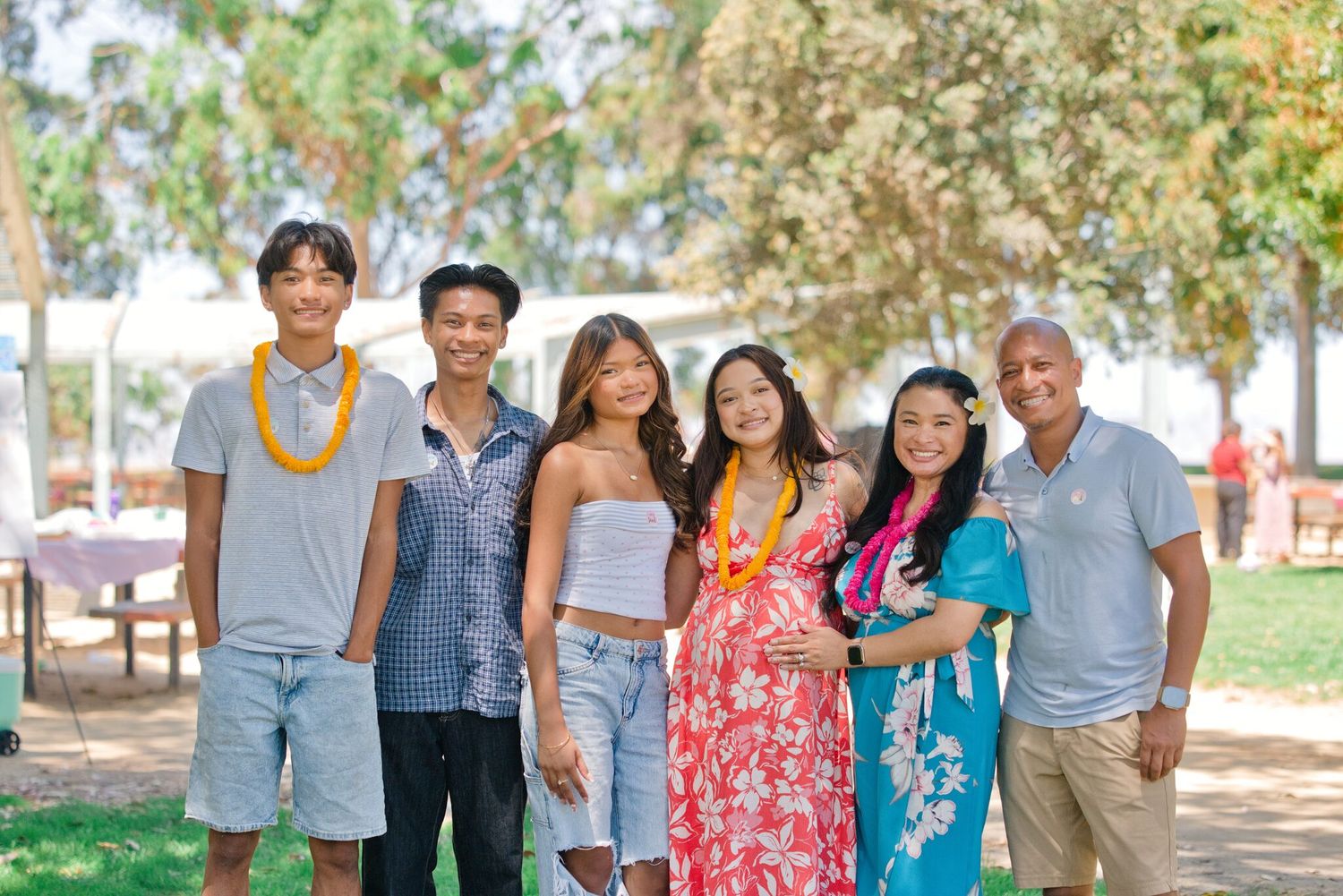 Family of six posing together outdoors at a sunny park gathering, some wearing floral leis and colorful Hawaiian attire.