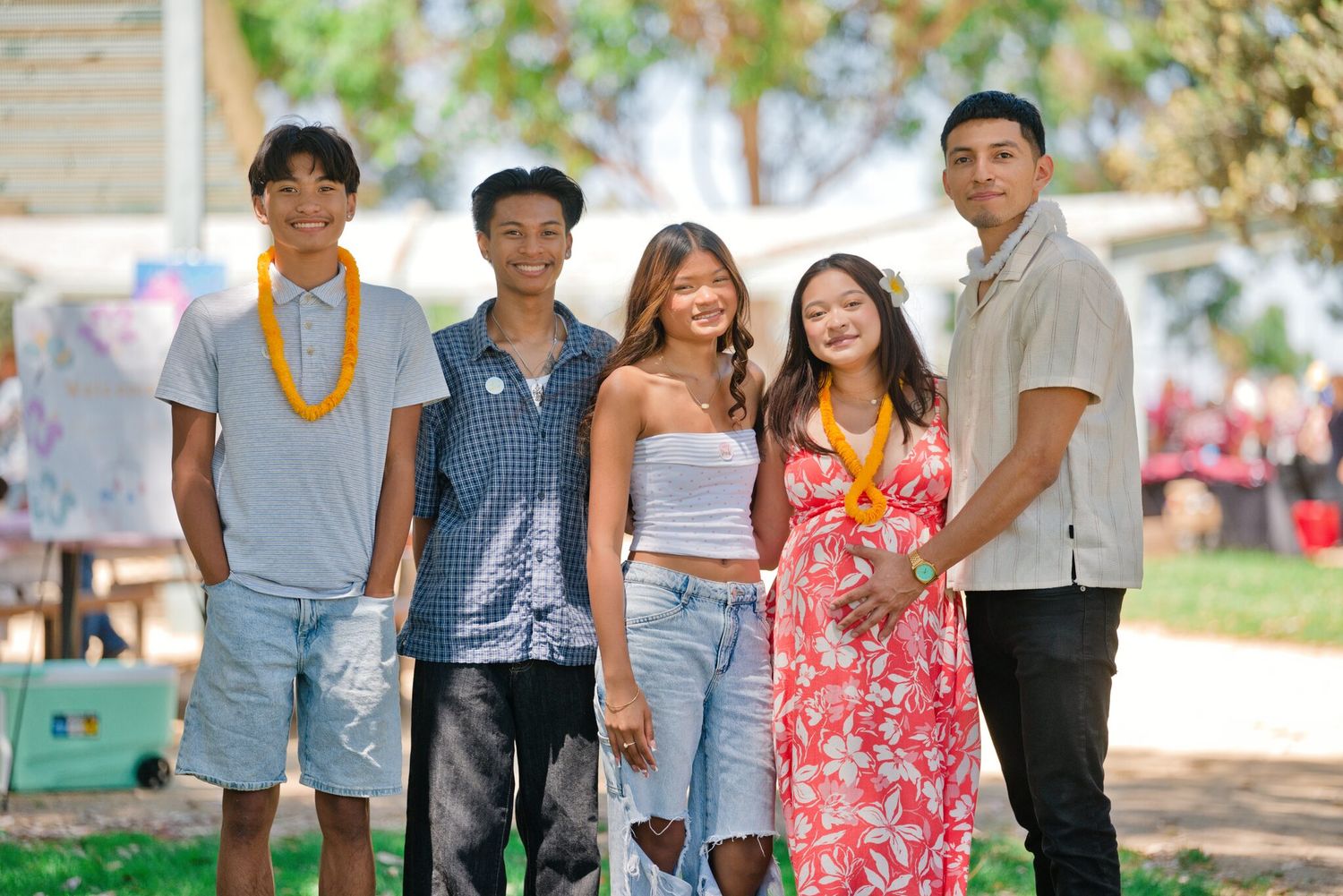 Group of five young people smiling outdoors at a sunny park event, wearing leis and casual summer outfits.