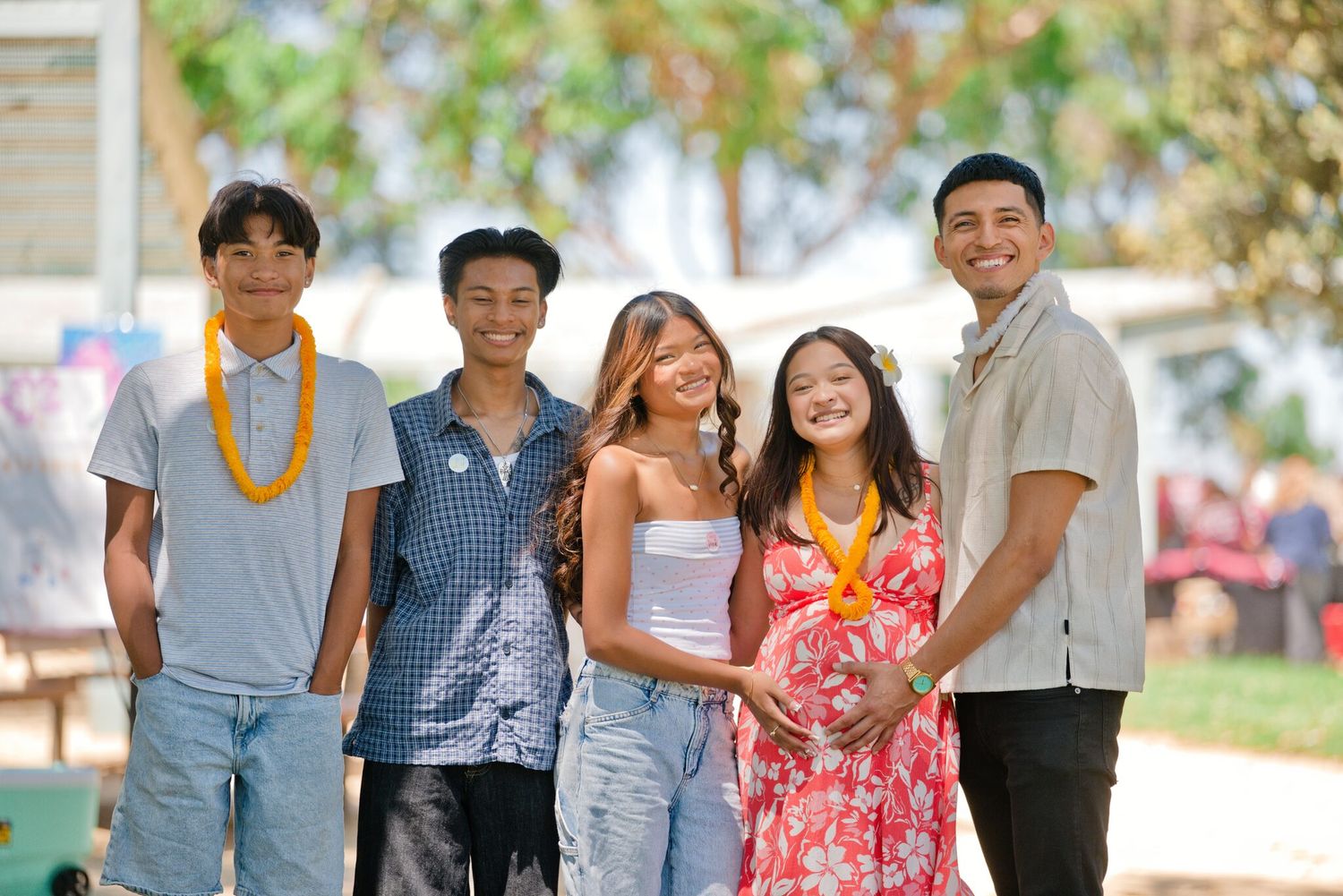 Five smiling young people posing together outdoors at a sunny gathering, some wearing floral leis and tropical attire.
