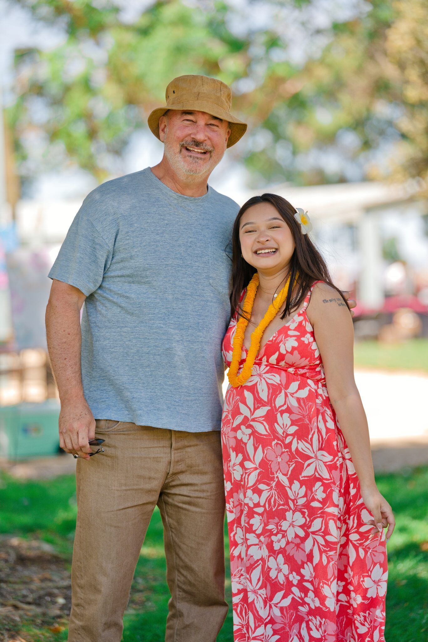 Smiling couple posing outdoors at a park, man in hat and gray shirt, woman in floral red dress with lei.