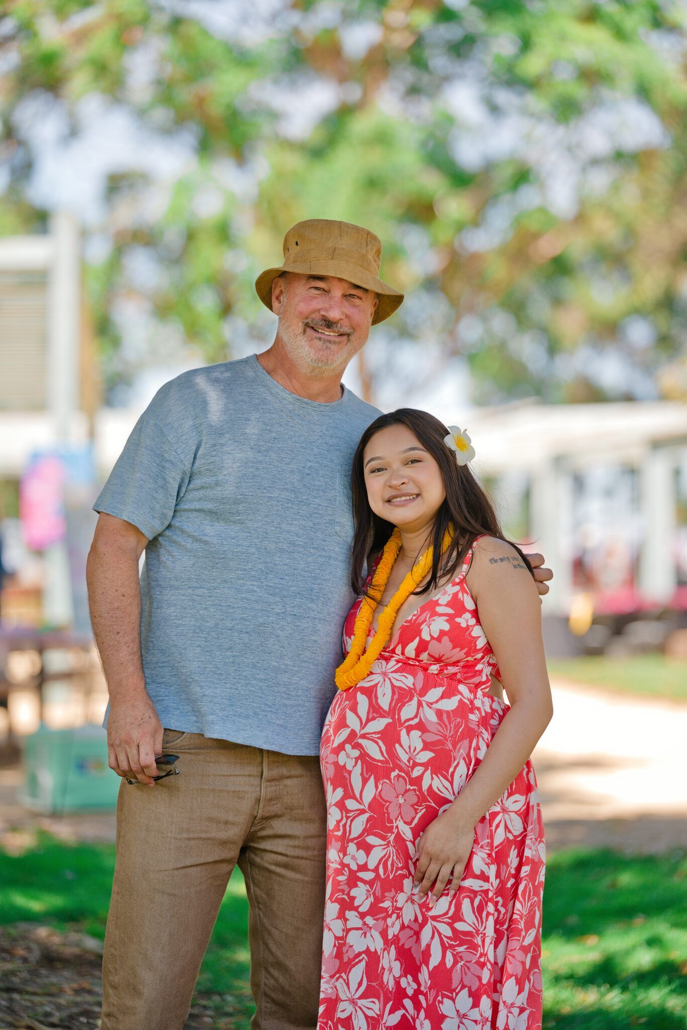 Happy couple embracing at sunny park, man wearing bucket hat, pregnant woman in red floral dress with yellow lei.