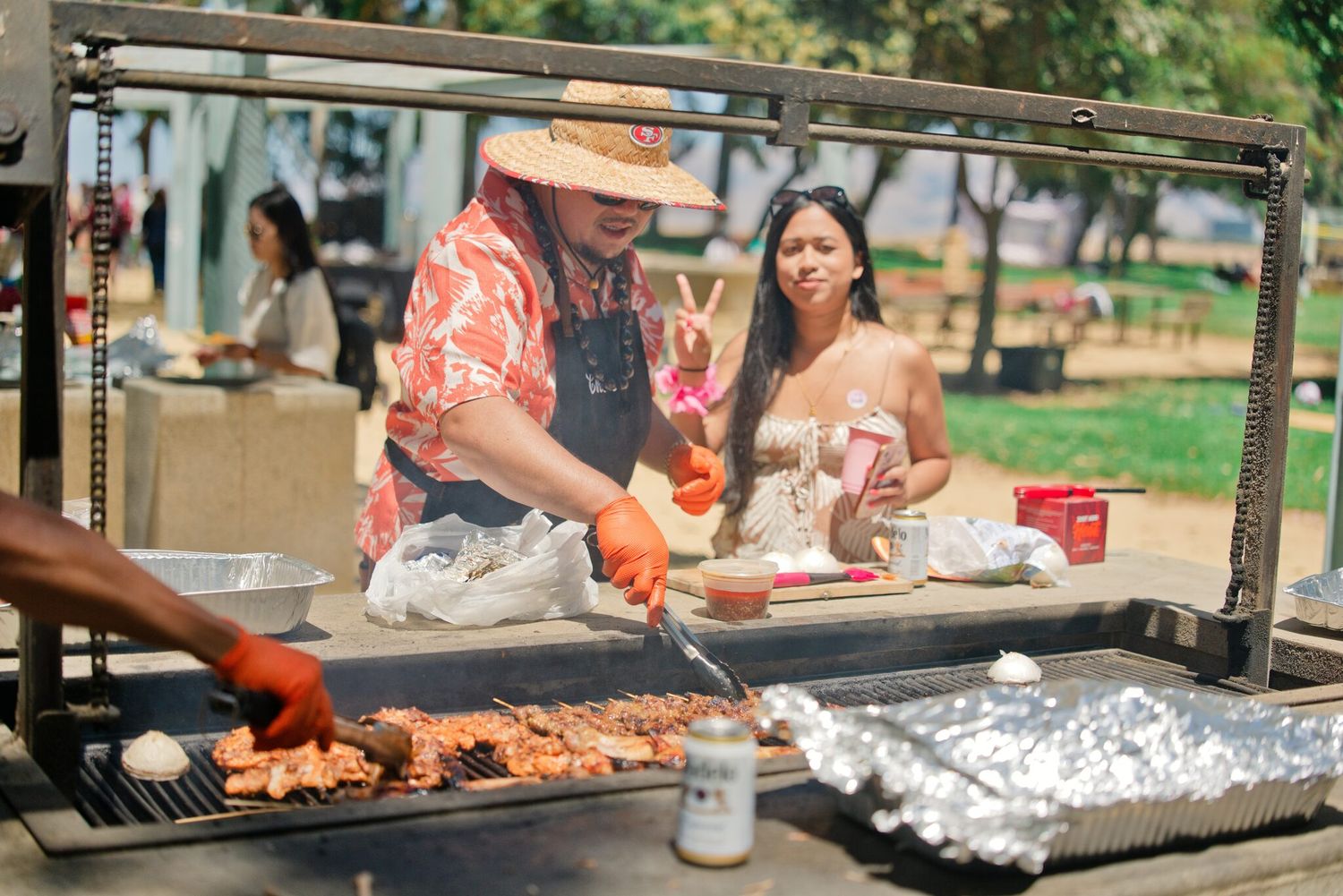 Man in floral shirt and hat grilling meat at outdoor BBQ event while woman watches in sunny park.