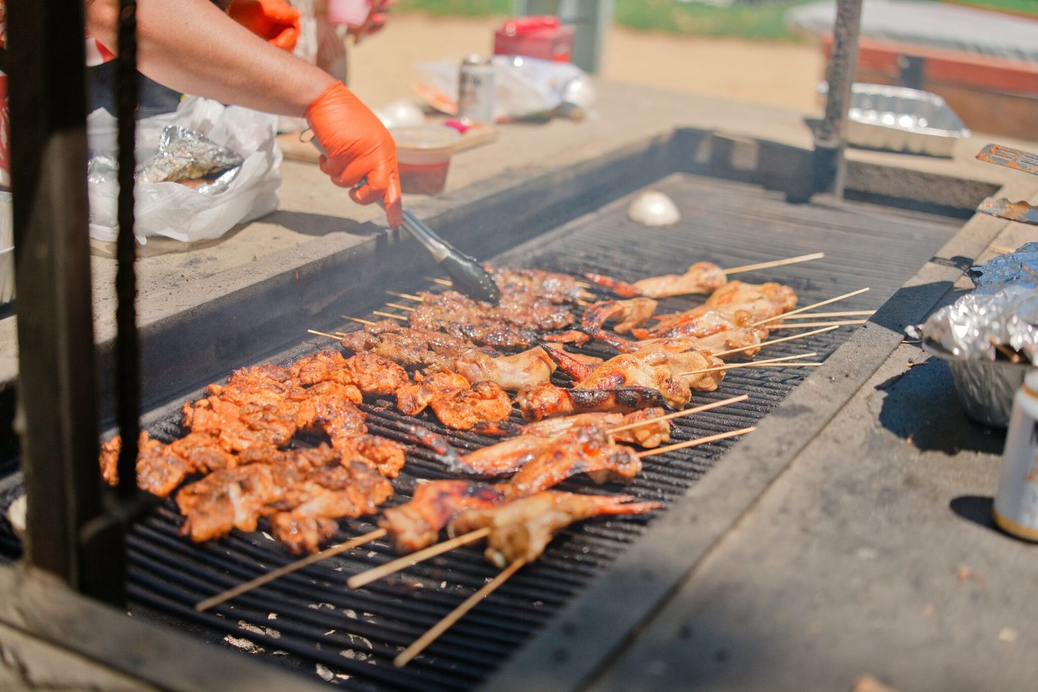 Person grilling marinated meat skewers on a large outdoor BBQ grill wearing orange gloves.