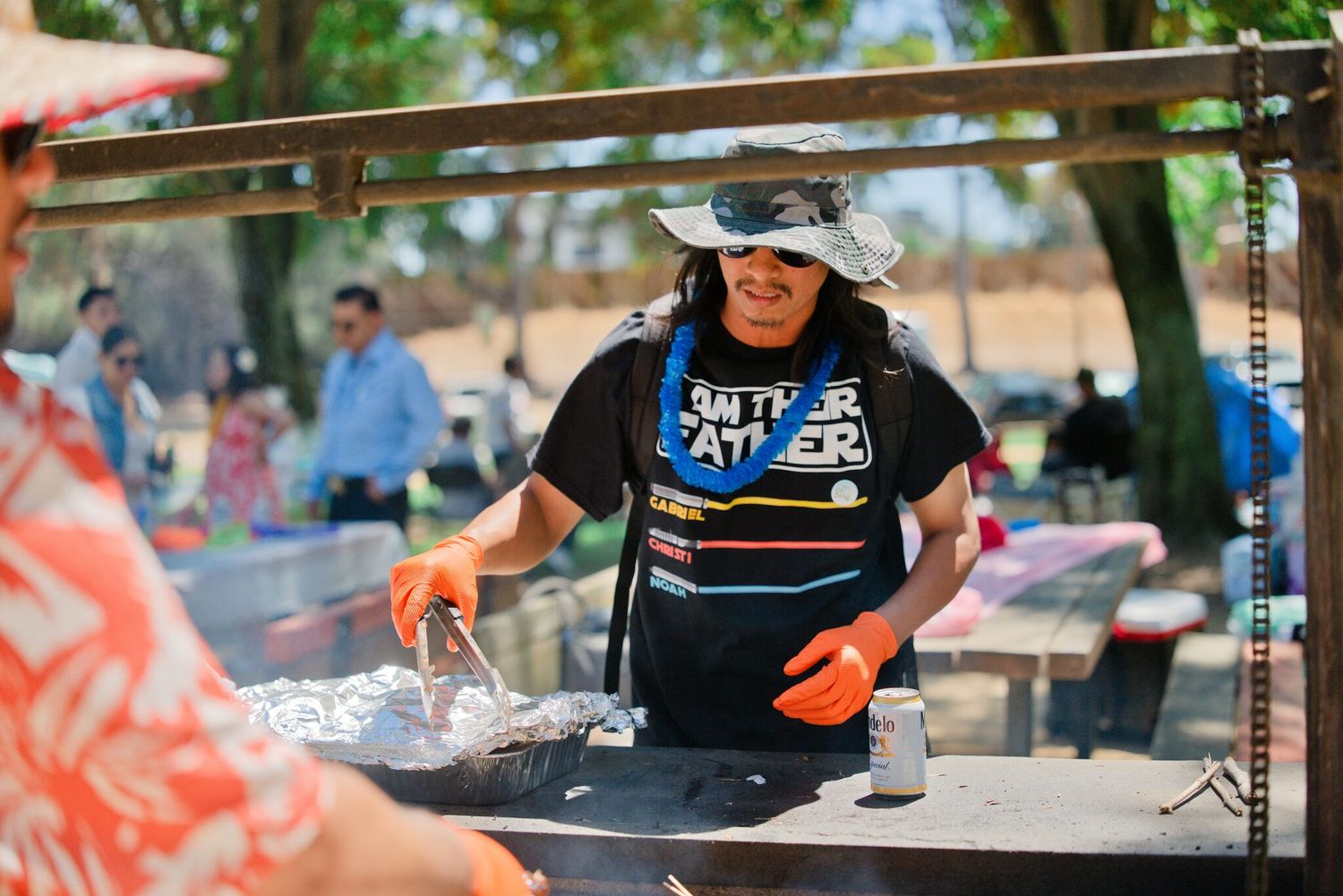 Person grilling food at outdoor barbecue wearing Black Panther shirt and orange gloves at park event.