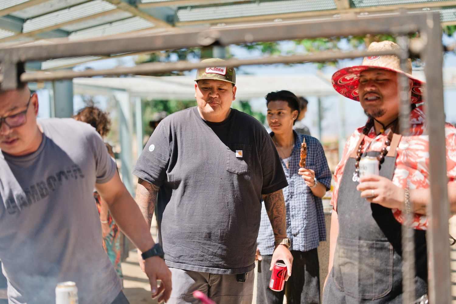 Group of people smiling and socializing outdoors at a sunny BBQ event, wearing casual clothes and hats.