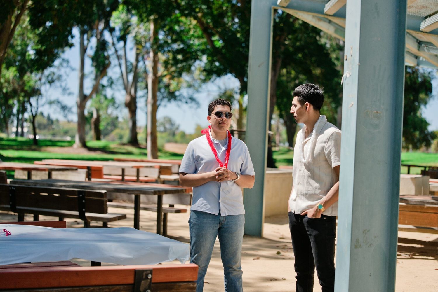 Two men having a casual conversation near a ping pong table in a shaded outdoor park area.