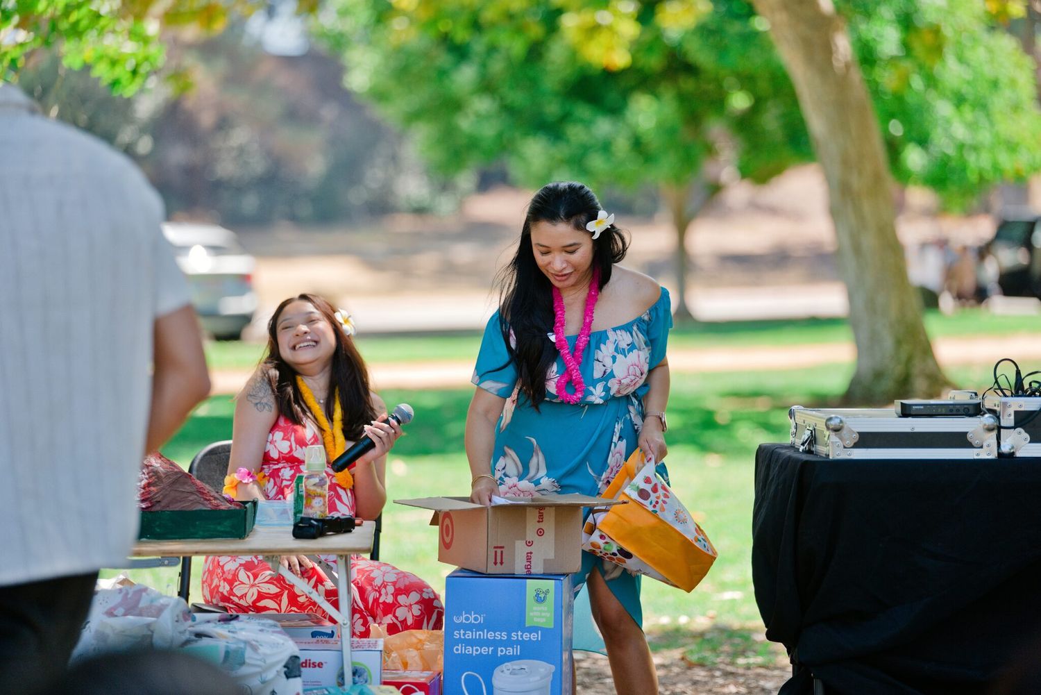 Two women laughing at outdoor event table under trees, wearing colorful Hawaiian attire with leis.
