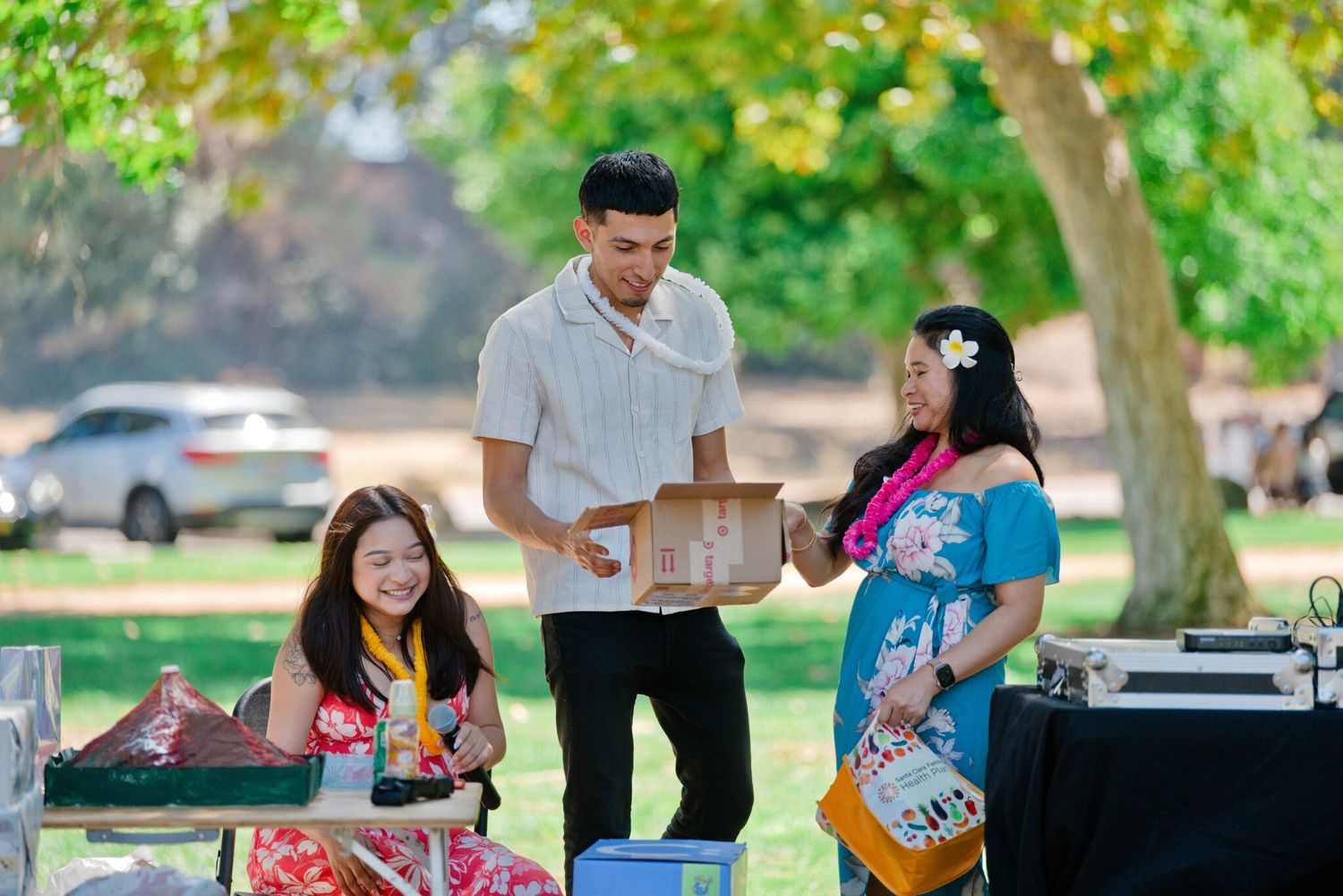 Young man presenting a wooden box to two women at an outdoor park event.