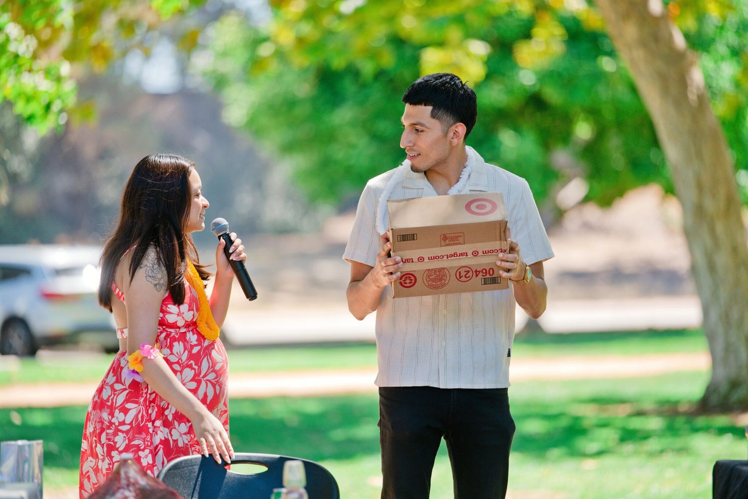 A man holds pizza boxes while a woman with a microphone interviews him outdoors in a sunny park.