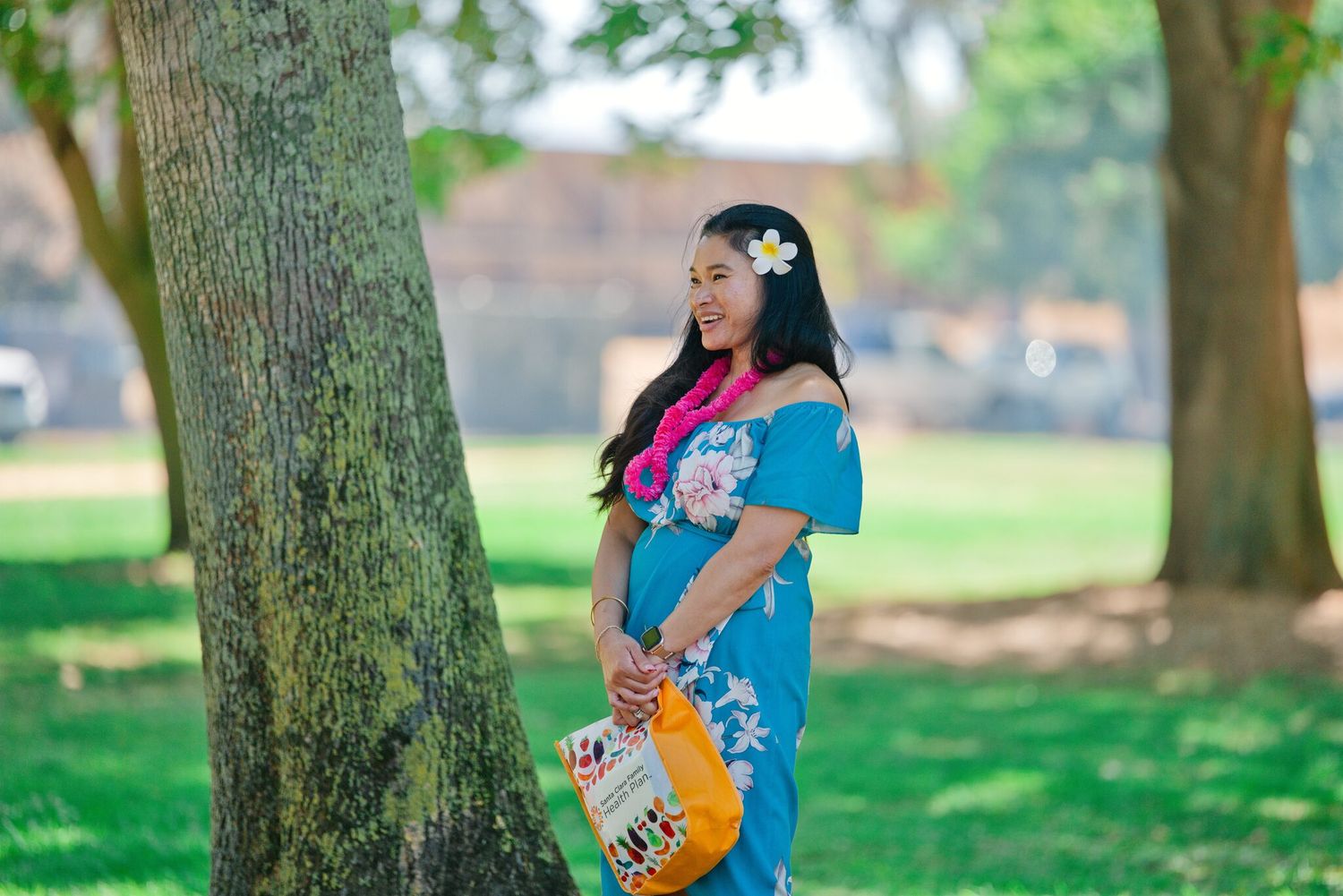Woman in blue floral dress with flower in hair standing by a tree in a green park.