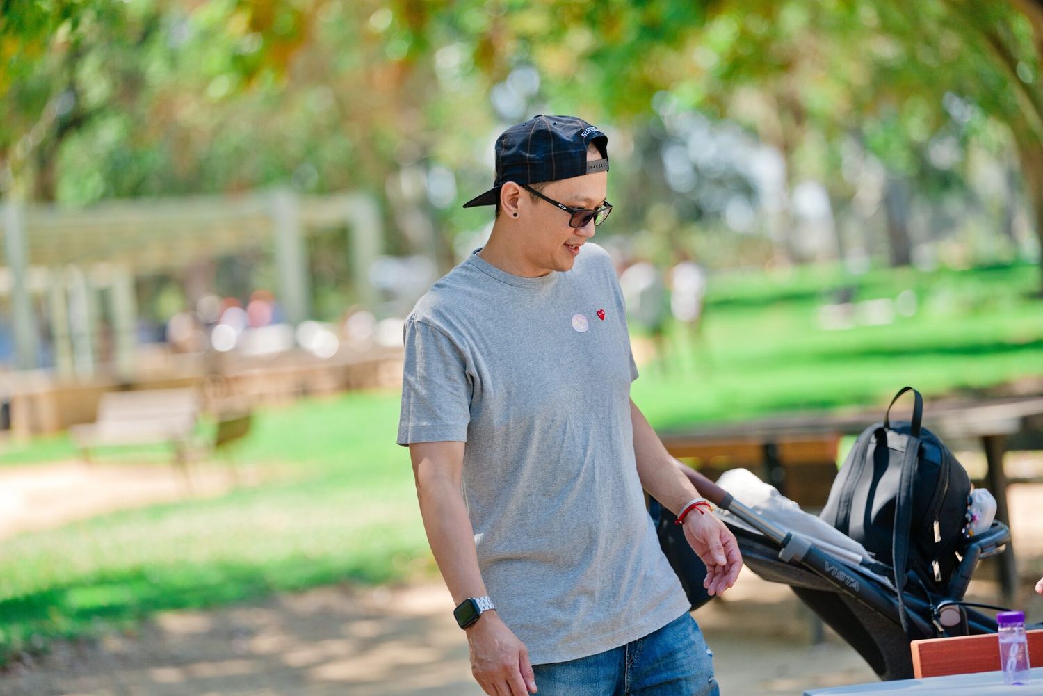 Man in gray t-shirt and baseball cap looking down at items on a table in a sunny park.