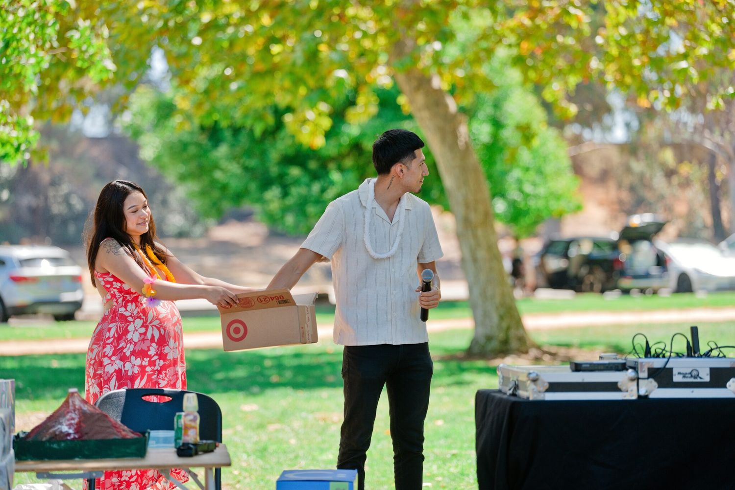 Young couple exchanging a box near DJ equipment under green trees at outdoor park gathering.