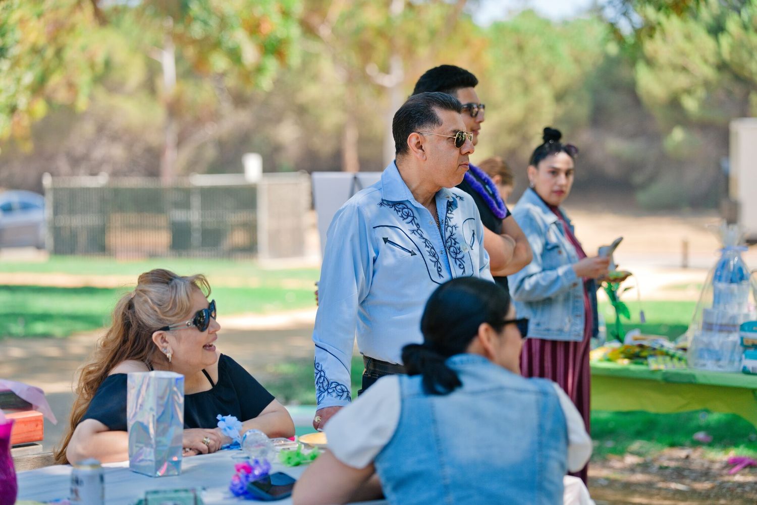 People gathered at an outdoor park picnic event with a man speaking into a microphone.