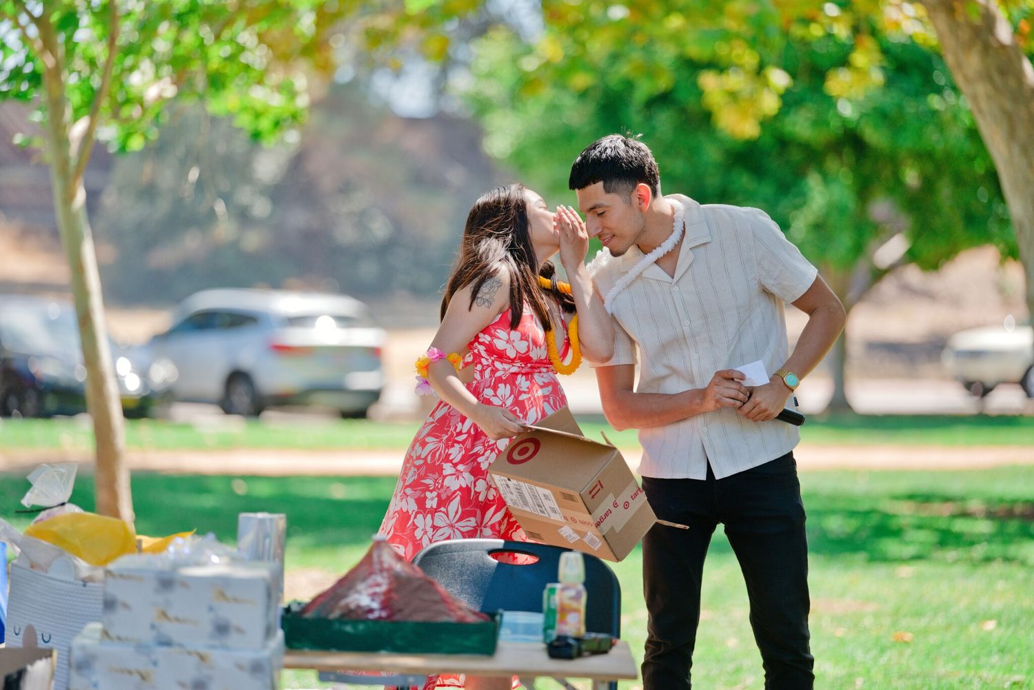 Young couple sharing a tender moment over gift table at outdoor baby shower in sunny park.