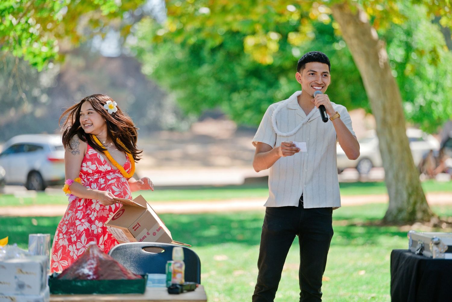 Young man speaking into microphone at outdoor park celebration while woman in red floral dress reacts joyfully.