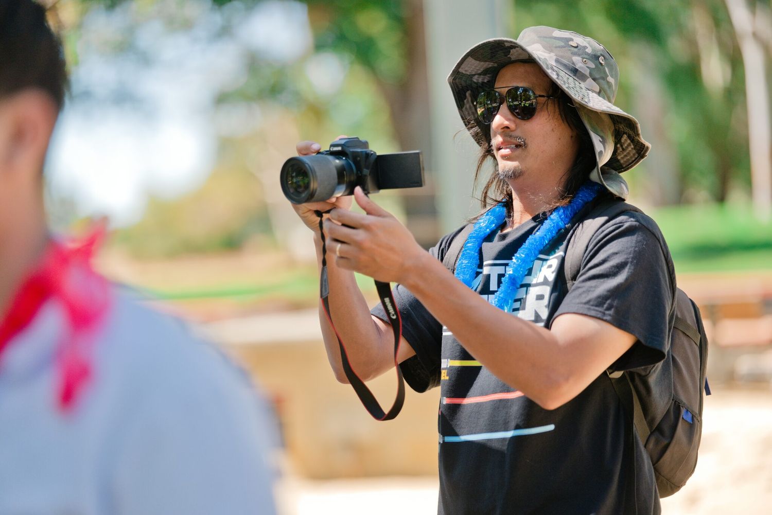 Photographer in bucket hat and sunglasses capturing photos with a DSLR camera at an outdoor event.