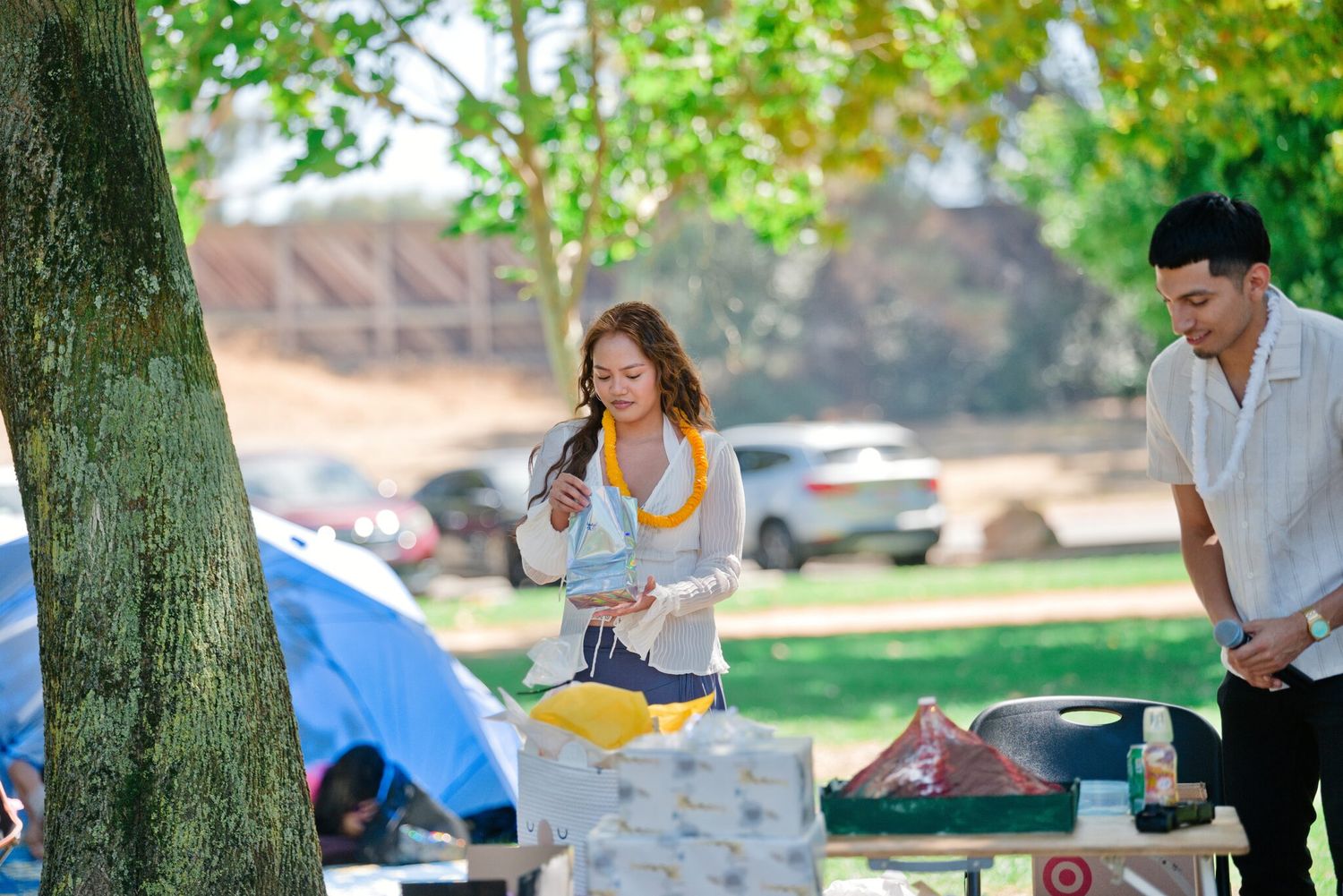 Pregnant woman in red floral dress with lei opens gifts at outdoor baby shower while man looks on.