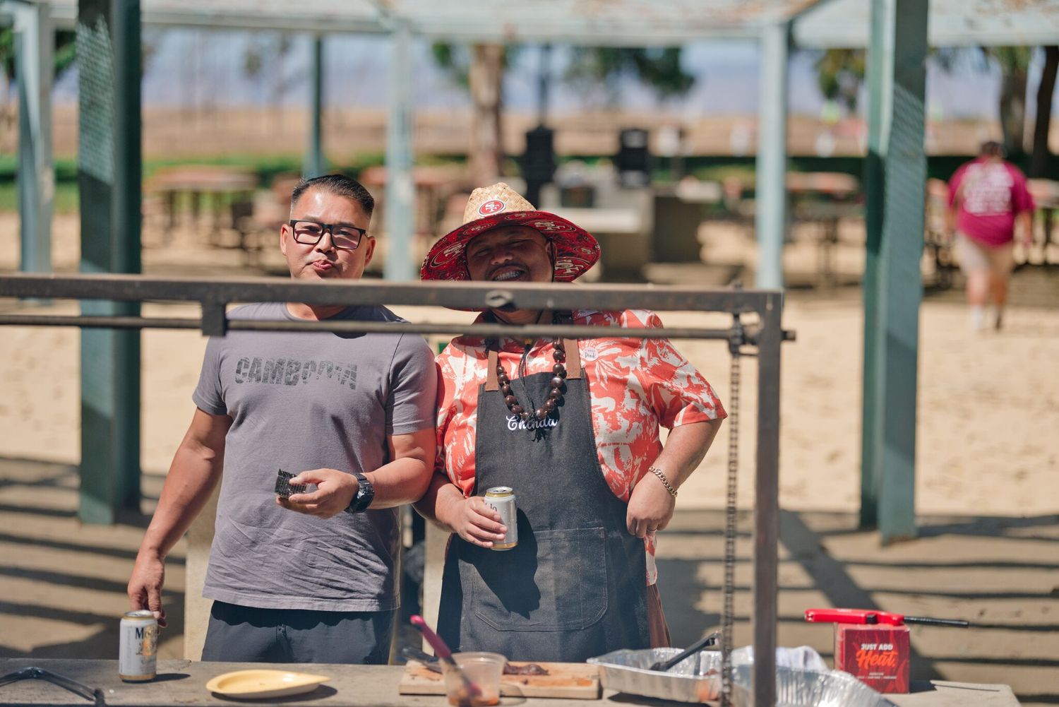 Two men standing at an outdoor grill cooking food at a sunny beachside barbecue event.