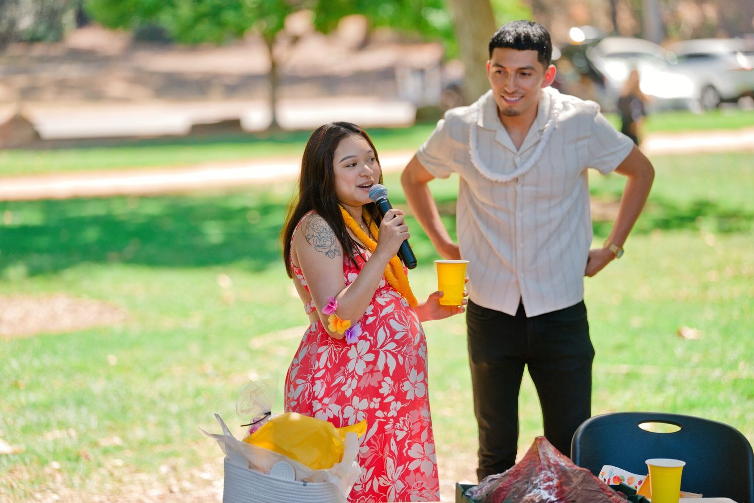 Woman in red floral dress speaks into microphone at outdoor park gathering while man holds yellow cup nearby.