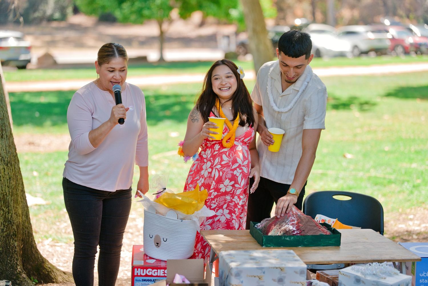 Three people at an outdoor picnic table opening baby shower gifts under trees in a park.
