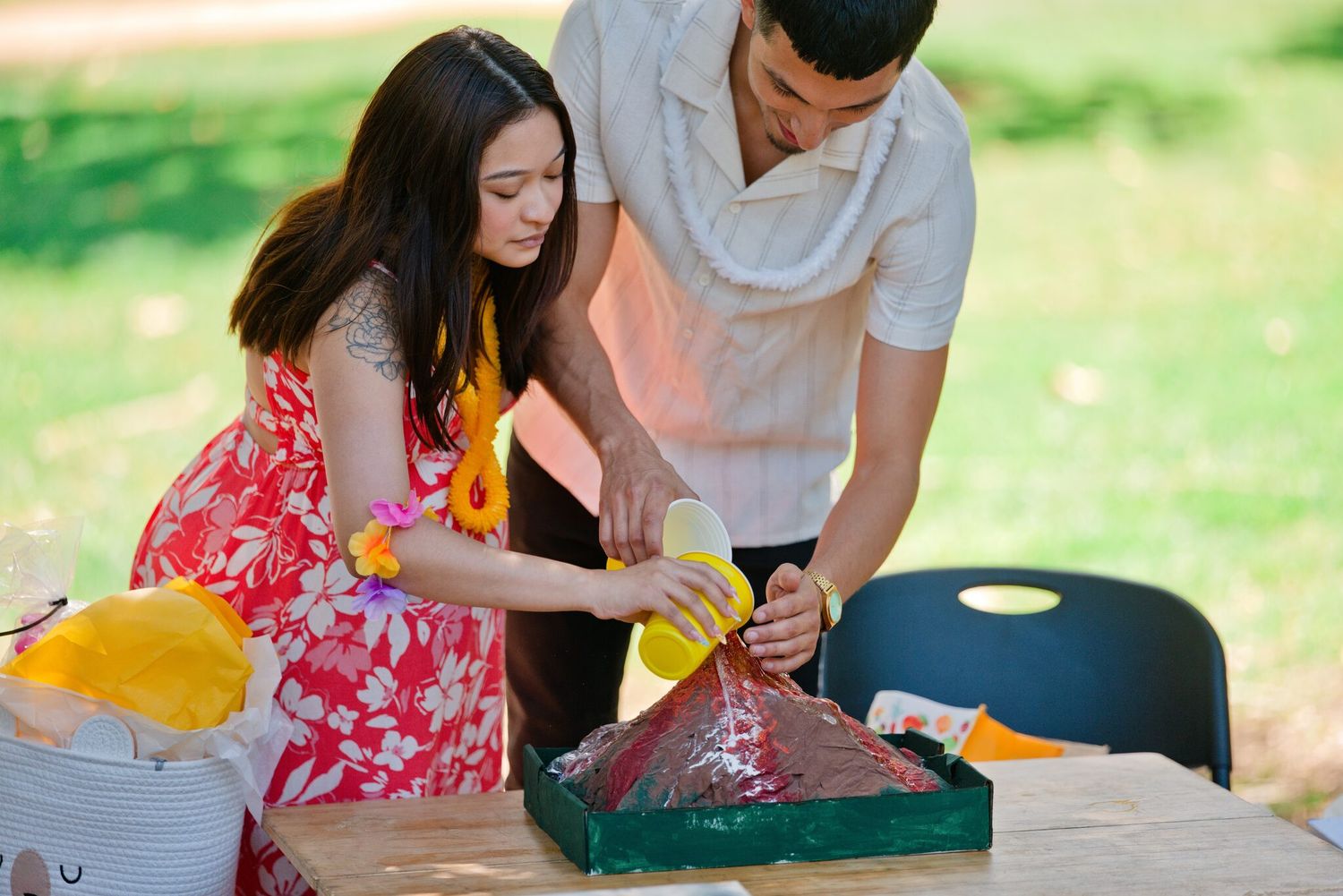 A couple works together outdoors at a table, pouring liquid over a colorful craft project in a sunny park.